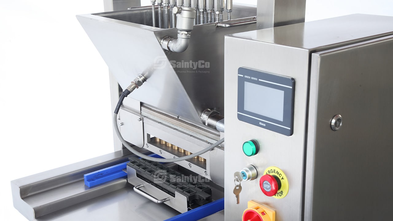 Close-up of a stainless steel tablet press machine used in pharmaceutical manufacturing, also ideal for gummy manufacturing equipment. The machine features a digital control panel, various buttons, a hopper filled with ingredients, and an output tray. The logo "SaintyCo" is displayed on the machine.