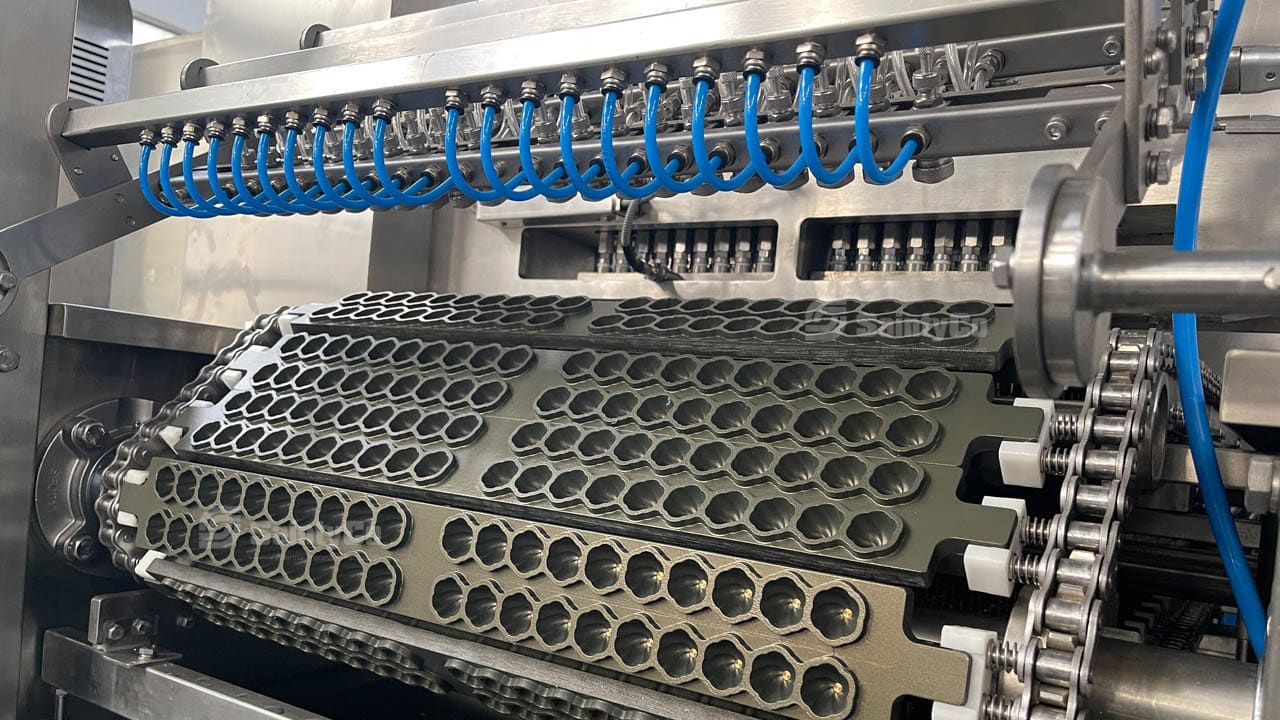 A close-up view of an industrial machine in a factory setting. The gummy making machine, possibly a SaintyCo model, features metal rollers with hexagonal molds, likely used for mass production. Blue tubing runs above the rollers, and the entire setup appears clean and well-maintained.