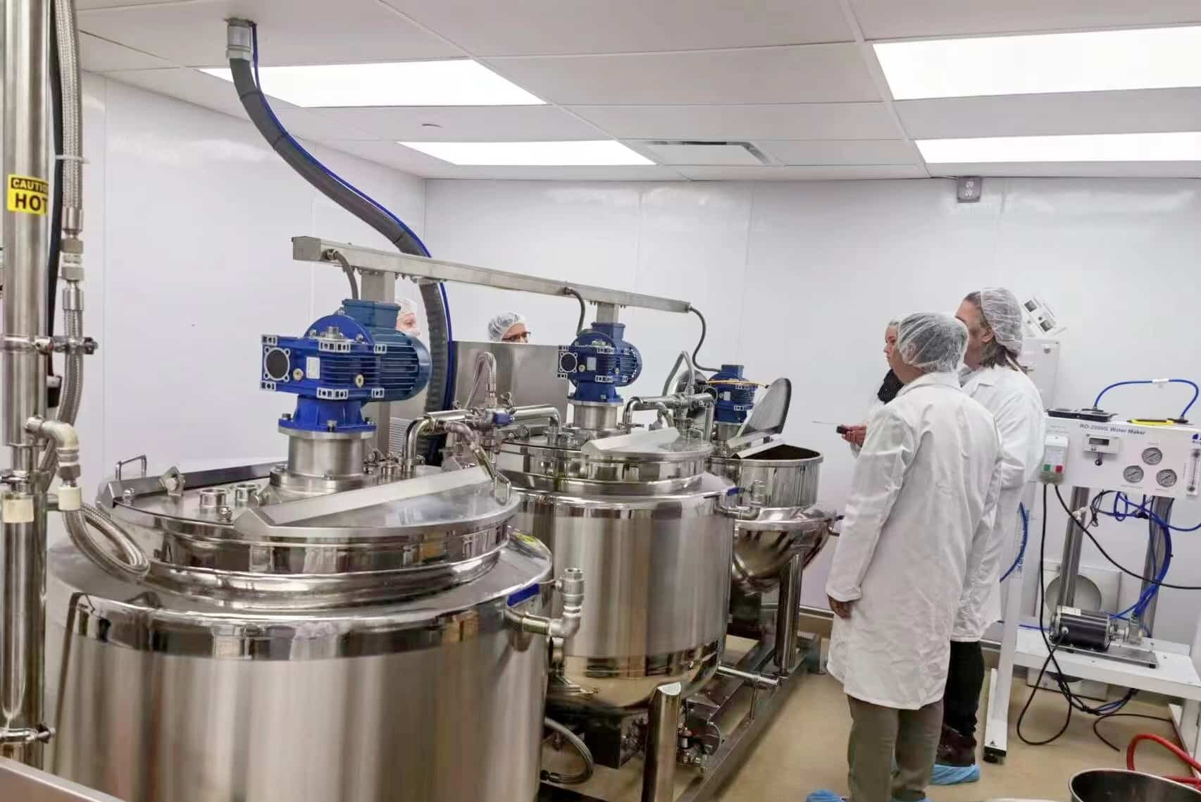 Several people in white lab coats and hairnets stand in a bright, clean laboratory, inspecting large stainless steel industrial mixing tanks with attached blue motors. Various pipes and control panels are visible in the background as they prepare to test a new gummy making machine by SaintyCo.