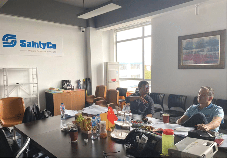 Two people sit at a conference table in a brightly lit SaintyCo office, deep in discussion. The table is cluttered with documents, drinks, snacks, and brochures on gummy manufacturing equipment. A sign on the wall reads "SaintyCo," and through the window, another building is visible next to a framed picture.