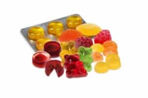 A collection of colorful gummy candies in various shapes, including bears, rounds, and shells, are displayed. The candies come in colors like red, green, yellow, and orange. Thanks to SaintyCo's advanced gummy manufacturing equipment, a blister pack of circular yellow gummies is also visible in the background.