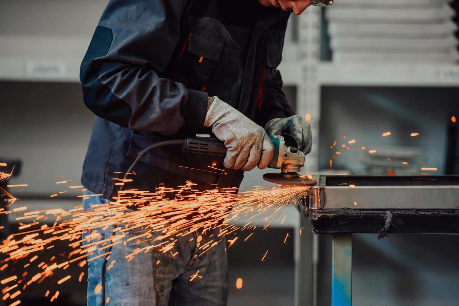 A person wearing protective gloves and a dark jacket uses an angle grinder on a metal piece, creating a shower of bright orange sparks. The background shows shelves with various items in a workshop setting, including tools for crafting gummy candies with precise gummy depostior equipment.