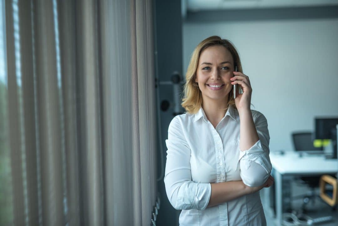 A woman with shoulder-length blonde hair is standing by windows with vertical blinds, smiling and talking on a phone. She is wearing a white button-up shirt with the sleeves rolled up. An office with desks, chairs, and a glimpse of gummy manufacturing equipment in the background is visible.