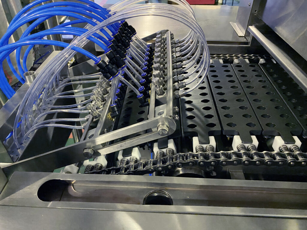 Close-up of an industrial gummy making machine of GummyGenix by SaintyCo. Multiple transparent tubes connected to valves and nozzles are positioned above a conveyor belt. The setup consists of metal components and chains, indicating complex mechanical parts used for automated gummy filling processes in the manufacturing industry.