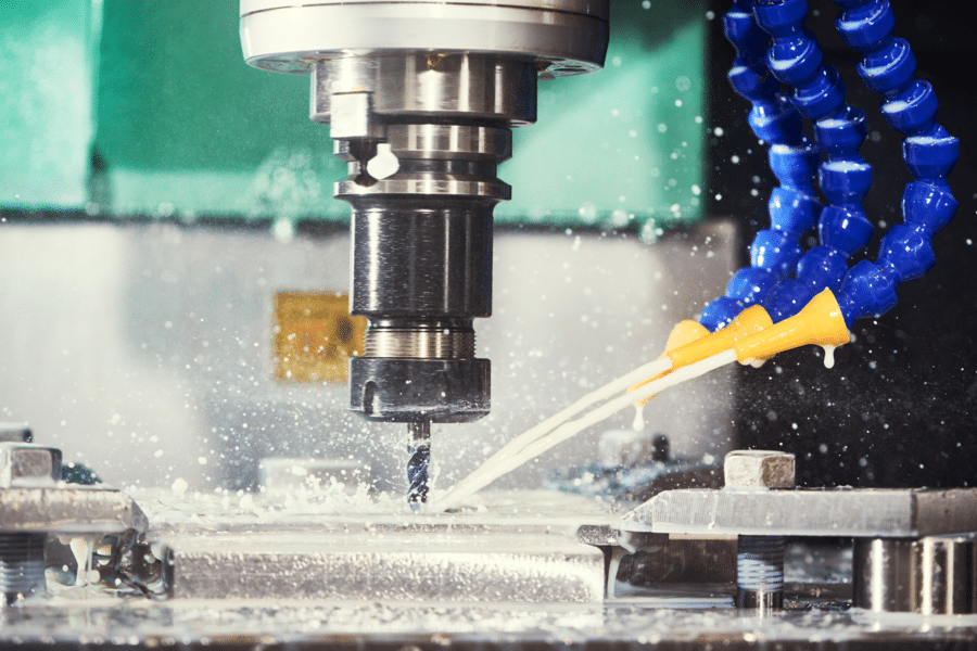 Close-up of a CNC milling machine in action. The machine's spindle is drilling into a metal surface while a coolant, delivered through blue flexible tubing, sprays onto the tool and workpiece, reducing heat and ensuring smooth operation. SaintyCo's precision is evident in every detail of the process.