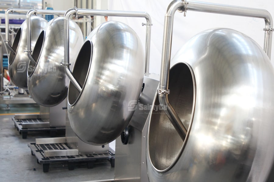 Four large, stainless steel spherical machines, likely industrial coating pans, are positioned in a row in a factory setting. These gummy manufacturing equipment pieces are shiny and metallic, with a hollow part facing forward. The background shows other industrial equipment and a pallet.