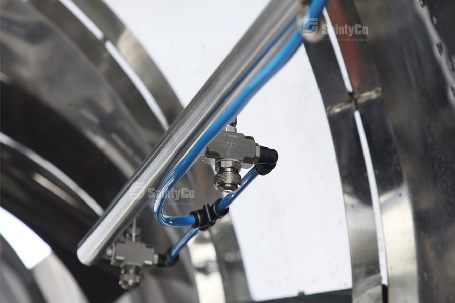 Close-up of industrial machinery featuring metal piping and connectors. Blue cables are connected to T-shaped metal junctions, mounted within a larger stainless steel structure. The equipment, which includes a gummy depositor, appears to be part of a manufacturing or processing system.