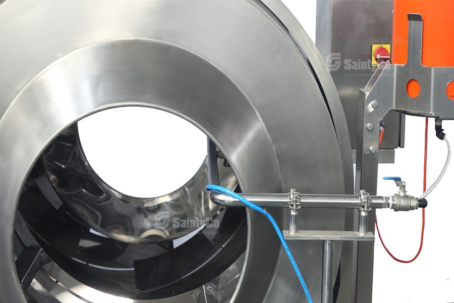 A close-up view of a large industrial stainless steel rotating drum with attached tubing and control equipment. The machinery has smooth, polished surfaces and some electronic components with wires visible, indicating a part of a gummy making machine in a manufacturing or processing plant.