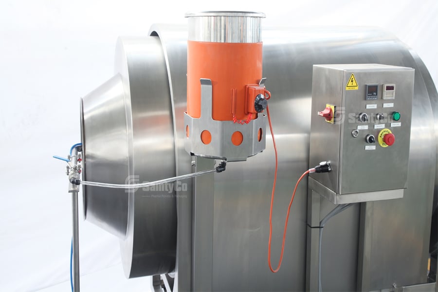 A close-up of a stainless steel gummy making machine by SaintyCo features a large rotating drum, an orange cylindrical attachment, and a control panel with buttons and switches on the right side. The setup is situated in a clean environment with a white background.