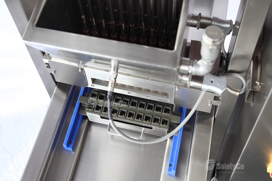 A close-up of industrial machinery, showing a stainless steel hopper feeding materials into a lower compartment. Blue guides are visible, with various mechanical parts and wiring. The setup appears to be part of a gummy manufacturing process or pharmaceutical production line.