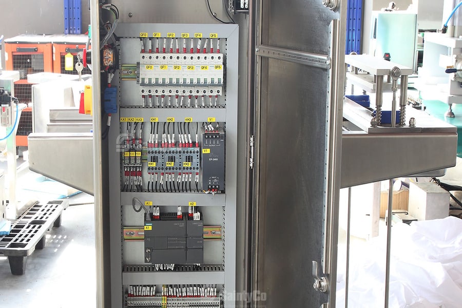 A metal electrical control panel with an open door reveals intricate wiring, switches, circuit breakers, and various components inside. Nearby, gummy manufacturing equipment and machinery are set up in a clean, well-lit environment ideal for production processes.