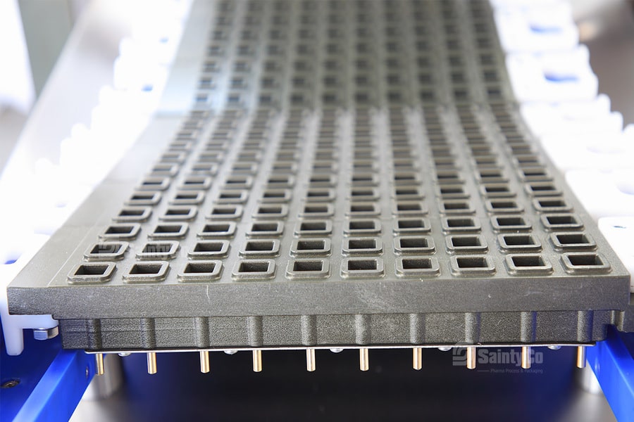 Close-up of a black industrial conveyor belt with a grid pattern. The belt, designed for the gummy depositor, has a series of small, square holes and rests on a metal structure with visible support pins underneath. The background is slightly blurred, emphasizing the texture and detail of the belt.