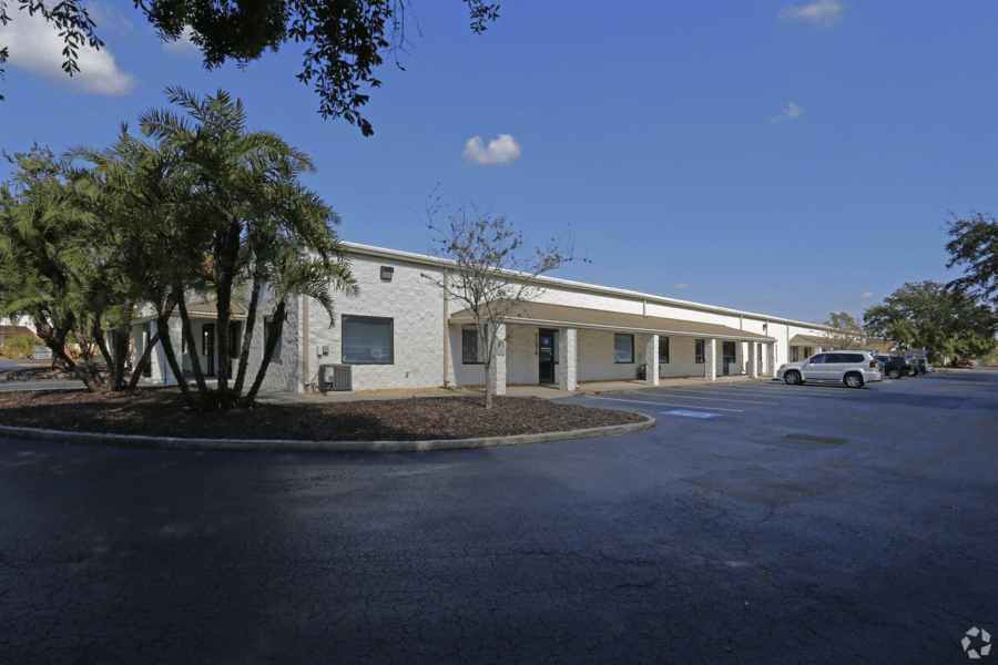 A one-story commercial building with a white facade and flat roof sits under a clear blue sky. Several cars are parked near the entrance, including a white SUV. Palm trees and other greenery are present around the building, which houses a state-of-the-art gummy making machine. The parking lot has empty spaces.