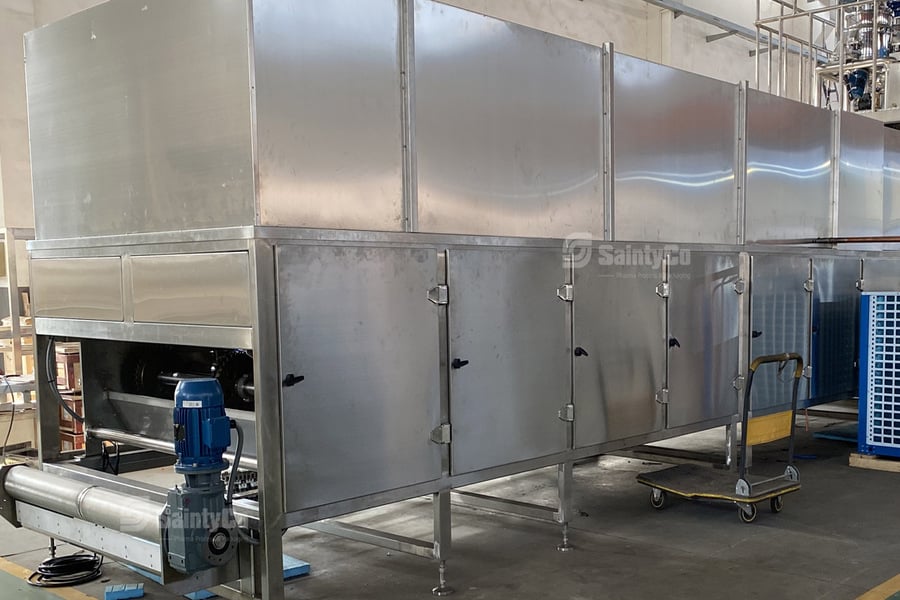 A large industrial stainless steel machine, likely a gummy depositor from SaintyCo, with multiple compartments for food processing or packaging, is seen in a factory setting. A blue motor is attached to the machine's lower section. Nearby, a blue pallet truck and some shelving are visible.