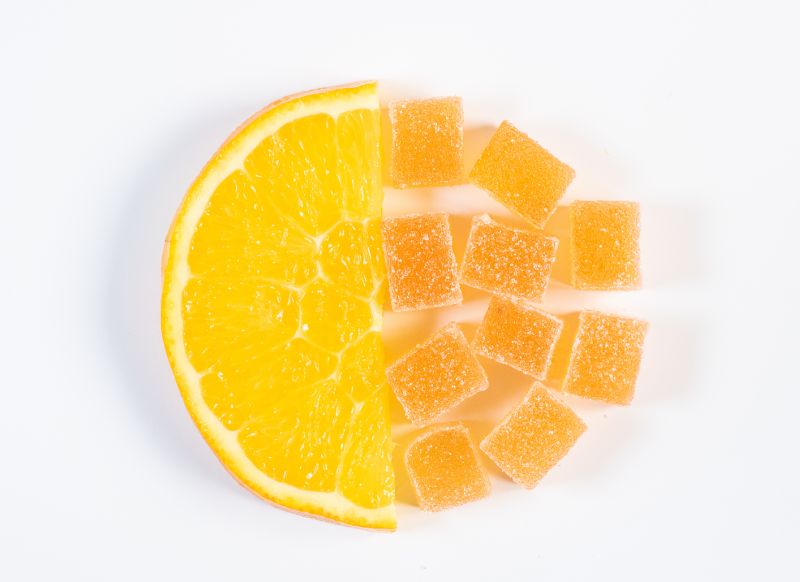 Half of an orange slice is placed on the left side of the image. Next to it, on the right side, are several orange-colored, sugar-coated jelly candies arranged in a grid pattern, mimicking the shape and size of the orange segments. All are set against a white background, courtesy of Gummygenix gummy making machine.