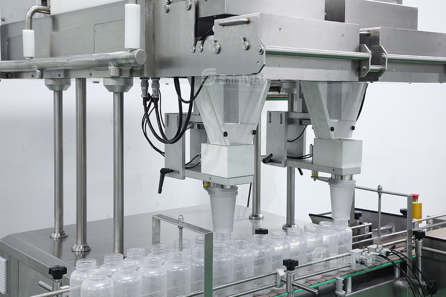 Industrial bottling equipment in a factory setting, with a machine funneling liquid into transparent plastic bottles on a conveyor belt. Stainless steel components and piping are visible, and the area is clean and well-lit. SaintyCo gummy manufacturing equipment seamlessly integrates with the process.