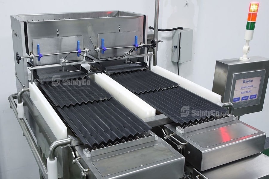 A close-up shot of a SaintyCo pharmaceutical blister packaging machine. The stainless steel device has black conveyor belts and blue valves. A control panel with buttons and a digital display is on the right side. In the background, there’s a vertical red and green indicator light, hinting at its gummy manufacturing capabilities.