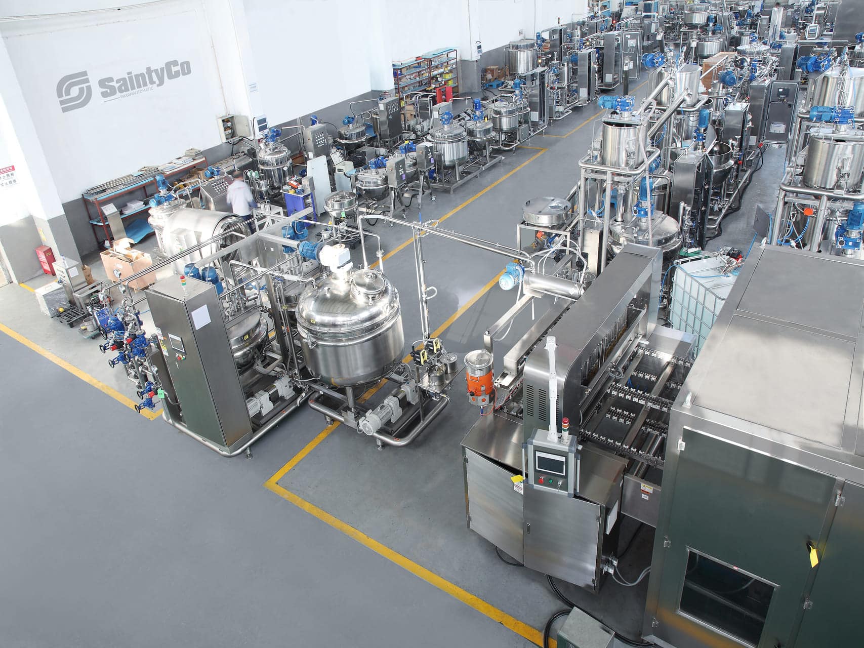 The image shows a modern, organized manufacturing facility. It features various industrial machines, large stainless steel tanks, and equipment arranged in rows. Several workers in white lab coats are seen operating the machinery. The floor is clean with marked pathways.