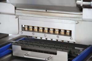 Close-up of a SaintyCo industrial machine dispensing liquid chocolate into a mold with multiple square cavities. The metallic structure, reminiscent of gummy manufacturing equipment, features several spouts aligned to fit the mold beneath, preparing chocolates for production.