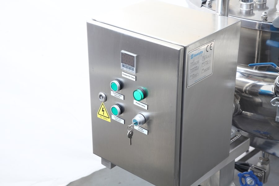 A stainless steel control panel with four green and blue buttons, a key slot, and a digital display on the front. The panel, branded by SaintyCo, is attached to gummy manufacturing equipment, partially visible, and includes safety and manufacturer labels.