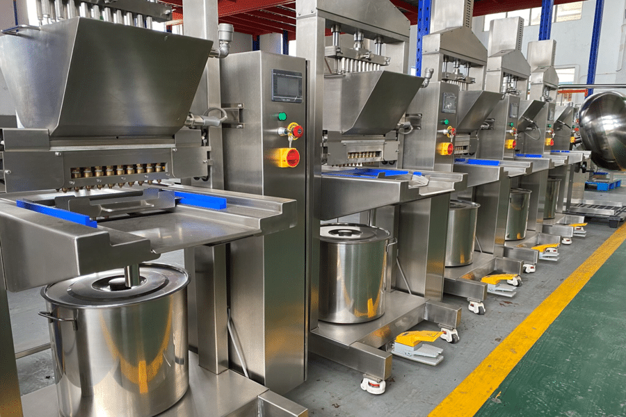 A row of stainless steel industrial food processing machines, including gummy manufacturing equipment with large hoppers and control panels, are lined up in a factory setting. Yellow foot pedals are at the base of each machine, which also has metal containers positioned underneath.