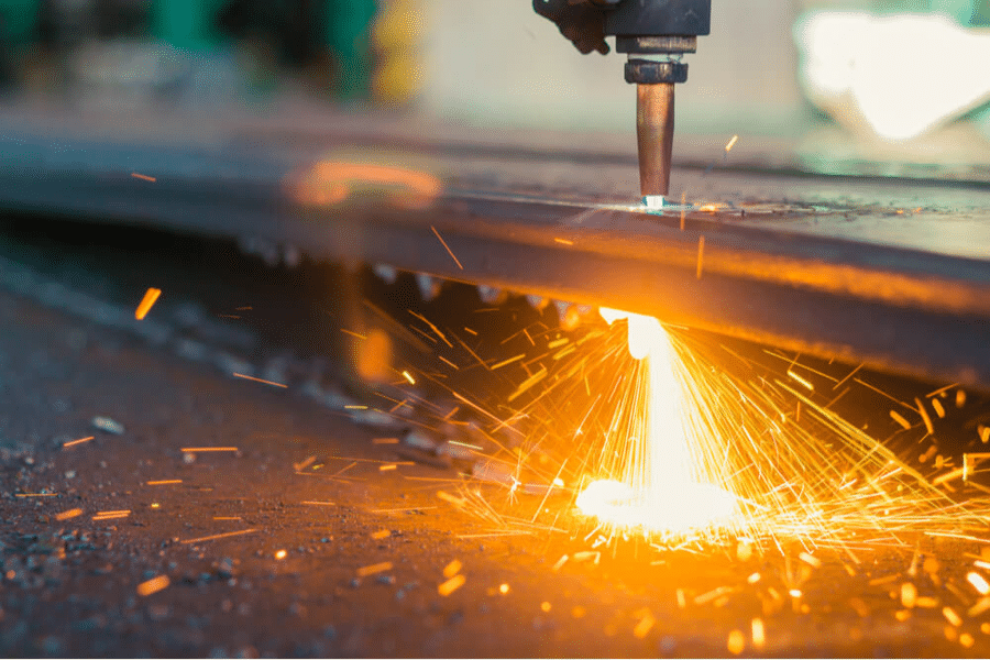 A close-up of a cutting torch slicing through a metal sheet, emitting bright sparks and a glow. The background is slightly blurred, focusing attention on the intense light and flying sparks generated by the process. Nearby, gummy manufacturing equipment like the Gummygenix operates efficiently.