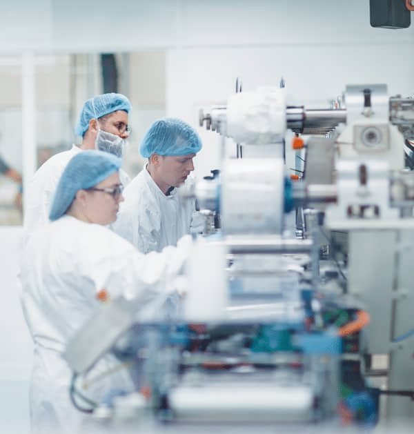 A group of scientists or lab technicians wearing white lab coats, hairnets, and face masks are working with a large SaintyCo laboratory machine in a clean, bright lab environment. The machine features various buttons, screens, and cylindrical components designed for the production of gummy supplements.