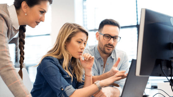 Three colleagues collaborate in an office, focused on a computer screen. A woman with a braid stands, while a woman in a denim shirt and a man with glasses sit closely, deeply engaged in discussion about the new SaintyCo gummy making machine, illuminated by natural light from the window.