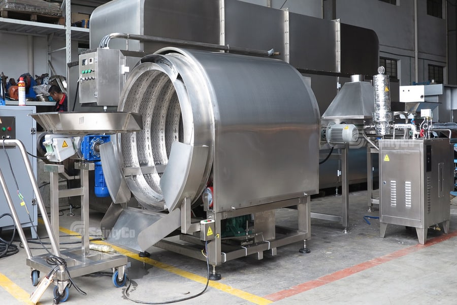 A stainless steel industrial rotary drum machine, integral to a gummy making operation, stands in a factory setting. The large cylindrical drum has various connections, controls, and safety equipment on its exterior. Surrounding the gummy making machine are various other pieces of related processing equipment.