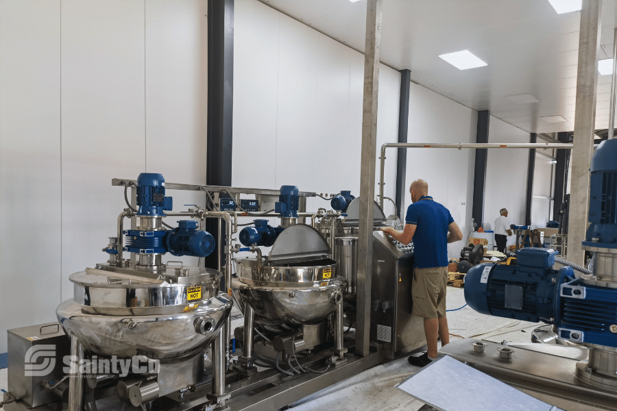 A person in a blue shirt operates machinery in a clean industrial facility with white walls. The room features large stainless steel mixing tanks and blue motors, part of a pharmaceutical or food processing plant. The "SaintyCo" logo is visible on one of the gummy making machines.