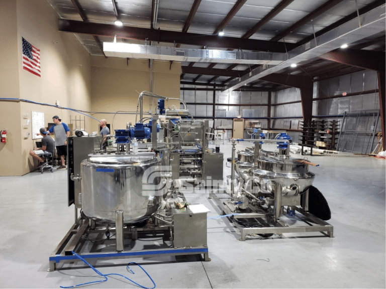 Factory floor with stainless steel industrial equipment, including tanks and specialized gummy manufacturing equipment. Five people are visible in the background, working and conversing near a table. The space has a high ceiling with exposed beams and an American flag on the wall.