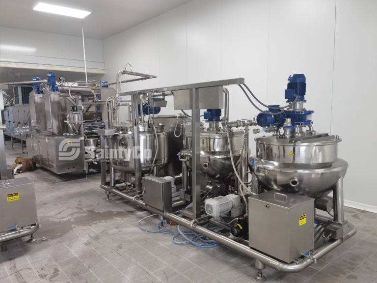 A stainless steel pharmaceutical or food processing machine with multiple interconnected tanks, pipes, and blue motorized mixers. The gummy manufacturing equipment is situated in a clean, industrial room with tiled flooring and white walls. The brand "SaintyCo" is visible on one part.