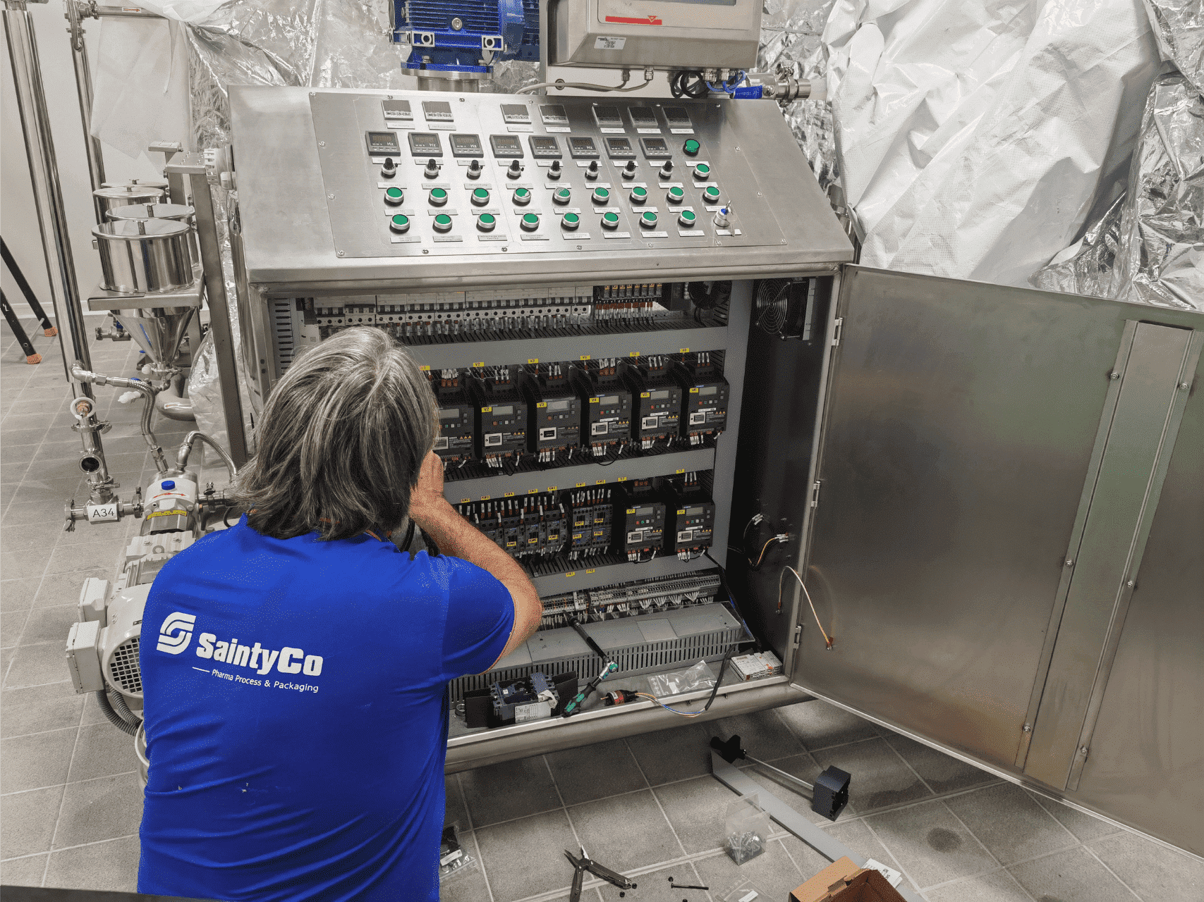 A person wearing a blue "SaintyCo" shirt works on an open industrial control panel with various switches, buttons, and wiring inside a metallic enclosure. Gummy manufacturing equipment surrounds the worker in a tiled room, and some tools and parts are on the floor.