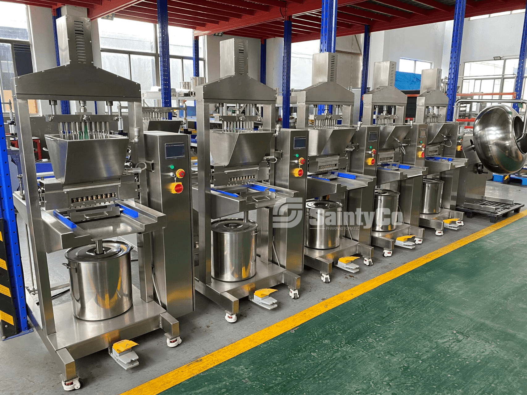 A row of industrial machines with stainless steel components, control panels, and blue trays is lined up in a factory setting. The machines, including a SaintyCo gummy depositor, are on a green floor, and the background includes red beams and equipment.