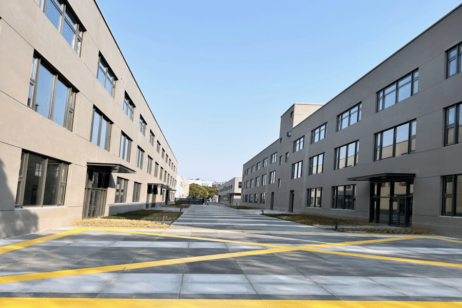 A wide pathway with yellow diagonal lines on the pavement runs between two modern, gray buildings housing state-of-the-art gummy manufacturing equipment. The clear blue sky above enhances the scene, and a few trees are visible in the background at the end of the pathway.