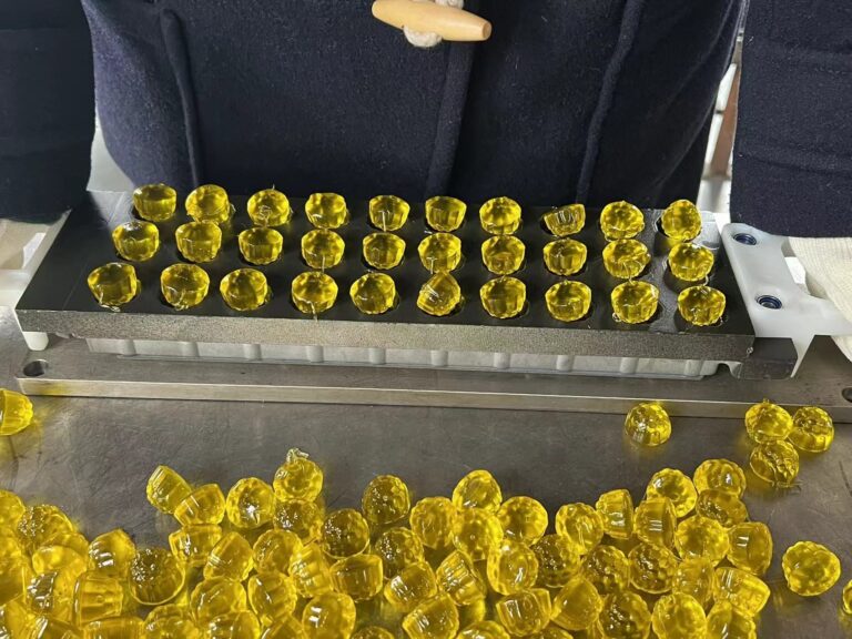 A person is preparing multiple yellow gummy candies using a SaintyCo candy mold tray. The tray holds a grid of filled cavities, and several finished GummyGenix candies are spread out on the stainless steel work surface below.