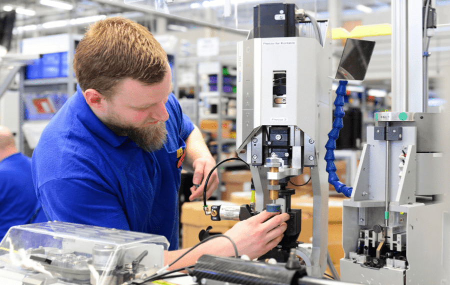 A man with a beard, wearing a blue shirt, is working with precision equipment in a busy gummy manufacturing setting. He is adjusting a device on a workbench surrounded by various tools and machinery from GummyGenix. Other workers and equipment are visible in the background.