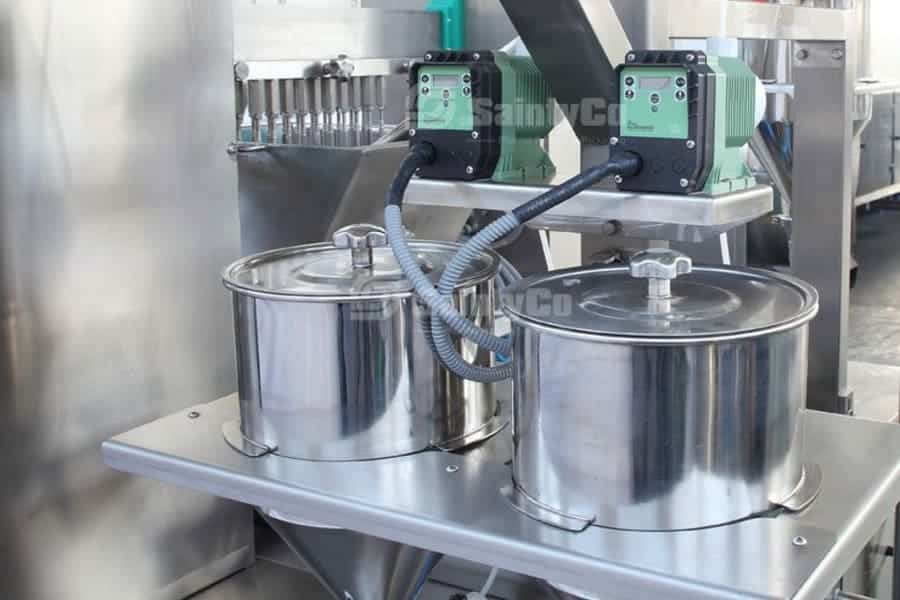 A close-up of a stainless steel SaintyCo industrial machine used for gummy making. Two large metal containers are positioned on a platform, connected by hoses to green gummy depositors mounted above. The background features additional metallic components and machinery.