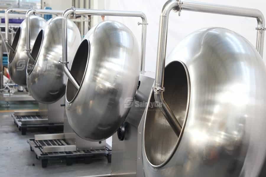 The image shows a row of large stainless steel industrial machines, including rotary coating drums and gummy-making machines used for coating chocolate on nuts or tablets in pharmaceutical settings. The machines are empty and placed in a clean, organized environment.