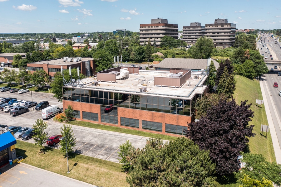 Aerial view of a two-story commercial building with a flat roof, glass facade, and brick exterior. Surrounded by trees and a parking lot, this Gummygenix location is near a highway with multiple office buildings and vehicles visible in the background.