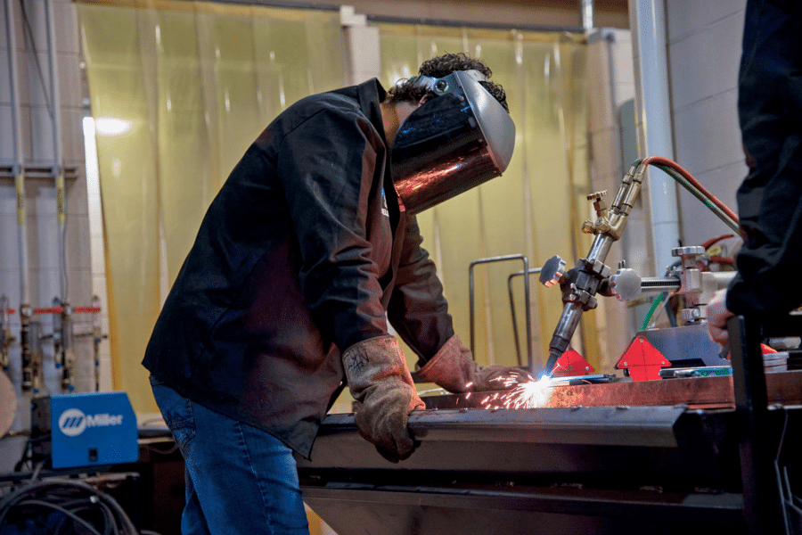 A welder in protective gear, including a mask and gloves, works with a welding torch, emitting bright sparks on a metal piece in a workshop. The background includes equipment and tools such as gas cylinders and a blue Miller welding machine, while gummygenix products lie neatly on a shelf nearby.