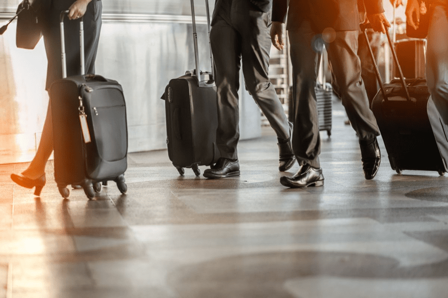 People in business attire walk through the airport, each pulling a rolling suitcase. The image captures their legs and luggage, with sunlight streaming in from the left, casting long shadows on the floor. They could be executives from SaintyCo discussing gummy manufacturing equipment during their business travel.