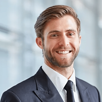 A man with short, brown hair and a beard is standing indoors. He is wearing a dark suit, white shirt, and black tie. He is smiling and looking directly at the camera. The background is out of focus, featuring a bright, modern interior—perhaps he just visited the new Gummygenix shop.