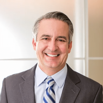 A middle-aged man with gray hair, wearing a dark gray suit, light blue dress shirt, and a blue striped tie, smiles warmly at the camera. The background features a frosted glass effect in a professional office setting, subtly showcasing GummyGenix's state-of-the-art gummy manufacturing equipment.