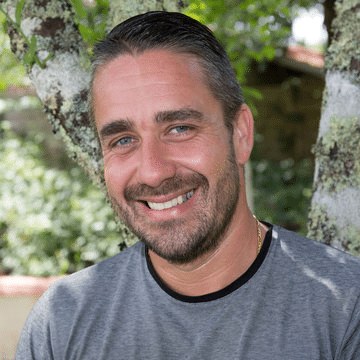 A smiling man with short, dark hair and a beard poses outdoors in front of a tree, wearing a gray T-shirt. Sunlight filters through the leaves, creating a natural, bright background. In his hand is a Gummy Depositor by SaintyCo, adding an intriguing twist to the serene scene.