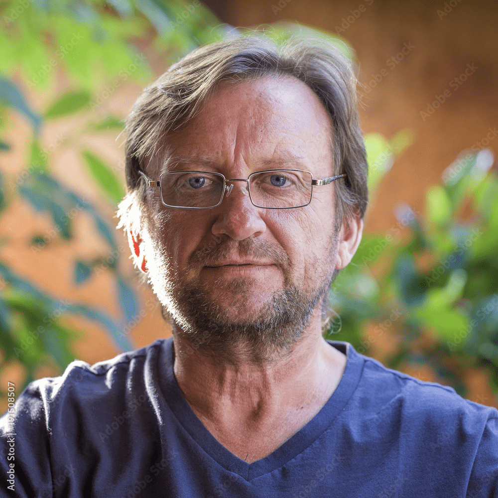 A middle-aged man with glasses, short brown hair, and a beard is looking directly at the camera. He is wearing a blue V-neck shirt and sitting outdoors with green foliage in the background. The natural lighting casts a warm glow on his face, making him appear as if he could be endorsing Gummygenix.