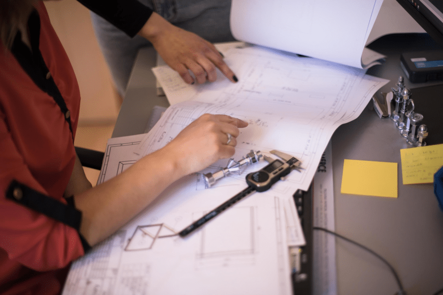 Two hands pointing at architectural blueprints spread out on a desk. A pair of calipers and a mechanical pencil rest atop the papers, alongside a gummy depositor sketch. The scene suggests a collaborative discussion or planning session, possibly in an office or workspace, with innovative ideas taking shape.