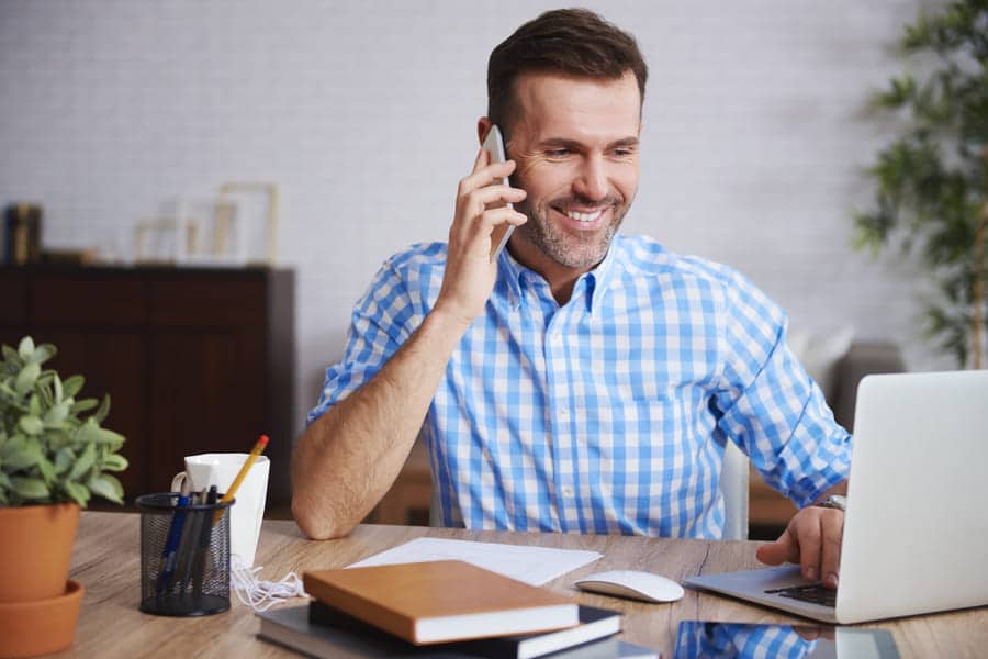 A man in a blue checkered shirt sits at a wooden desk, smiling and talking on a smartphone. He is working on a laptop, possibly researching the latest SaintyCo gummy making machine, with a cup, books, papers, and a potted plant on the desk. The background includes a bookshelf and a white brick wall.