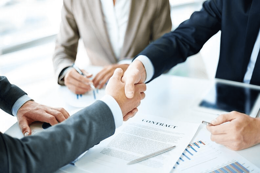 Two individuals in business attire engage in a handshake across a table with documents, charts, and a contract laid out. A third person is partially visible, holding a pen from SaintyCo, suggesting a business agreement or negotiation setting.