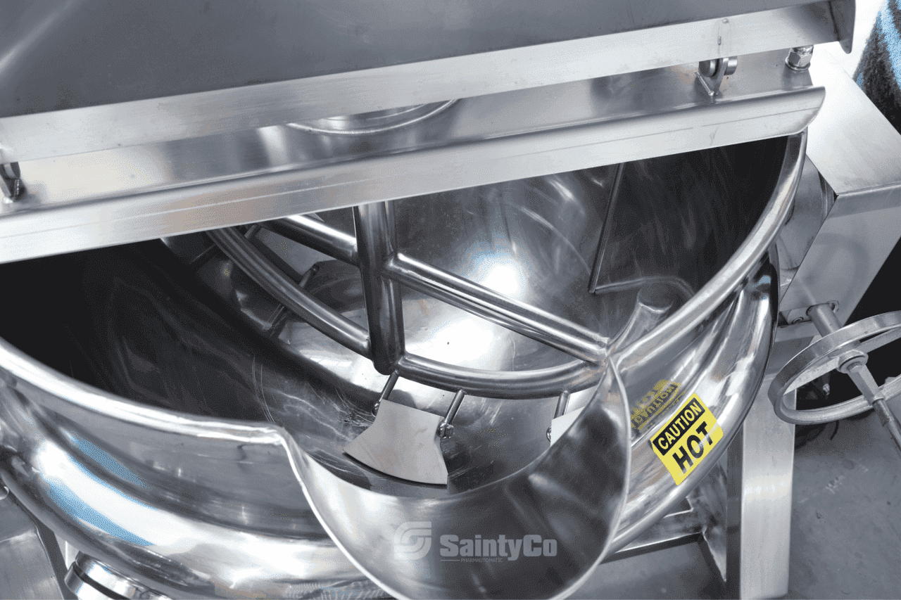 A close-up view of a large, industrial stainless steel mixing tank with a churn inside. The tank has a "Caution: Hot" warning label on it. The image prominently features the brand name "SaintyCo" at the bottom, illustrating its role in their advanced gummy making machine, Gummygenix.